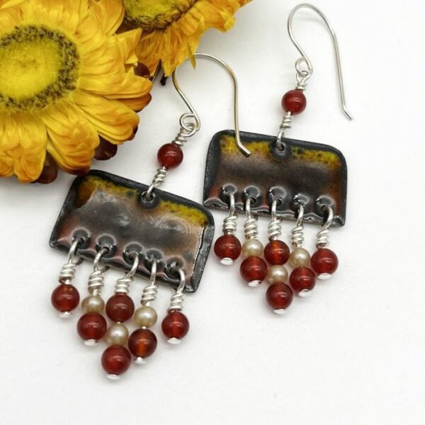 enamel drop earrings in shades of brown, red and yellow with red beads and warn coloured pearls suspended from the main oblong enamel piece. on silver ear wires