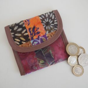 A coin purse made from a patchwork of cotton batik fabrics in autumnal colours, shown closed with a selection of coins