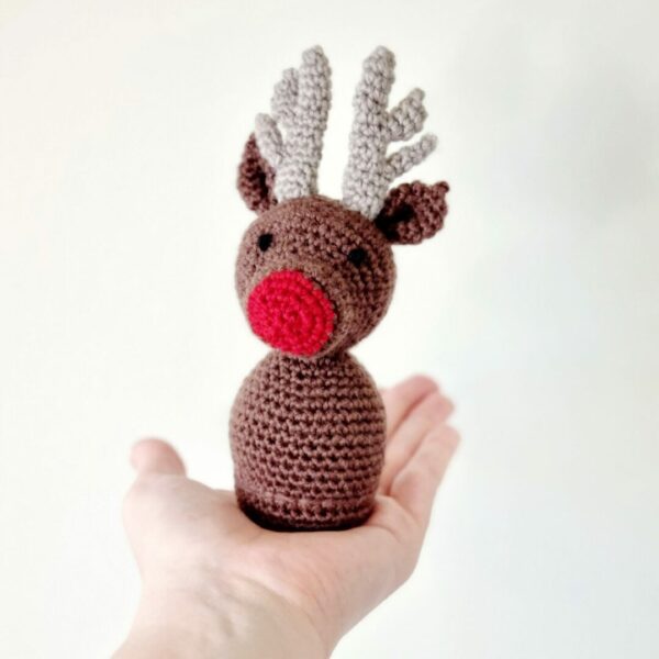 Small brown crochet reindeer soft toy sat in the palm of a hand on a white background