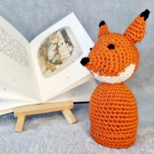 A crocheted orange fox soft toy sat next to a small open book