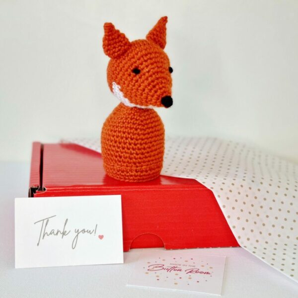 A orange fox soft toy sat on top of a red gift box, gold spotty tissue paper, a white card saying thank you and a square white business card.