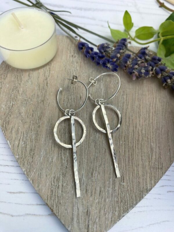 Silver circle and sticks attached to hoop earrings sitting on a wooden heart with candle and lavender