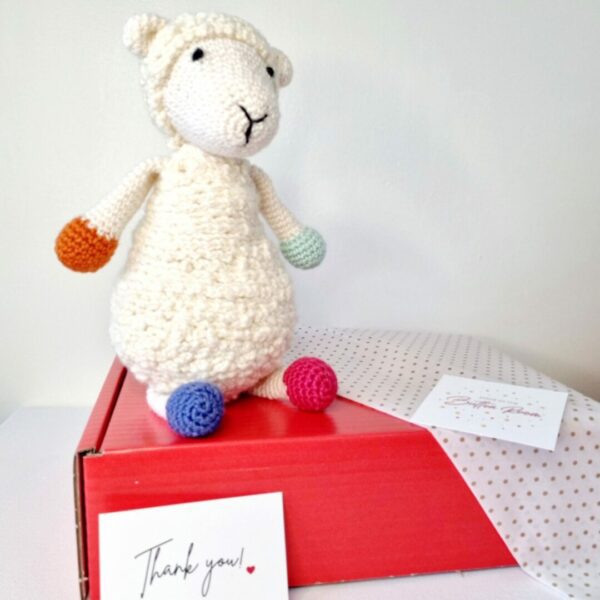 A large white crochet soft toy sheep sat on top of a red postage box with gold polka dot tissue paper, business card and thank you card.