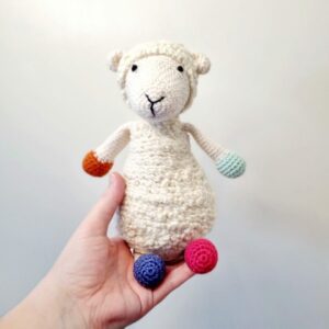 A hand reaching out whilst holding a large white crochet sheep soft toy with colourful toes.
