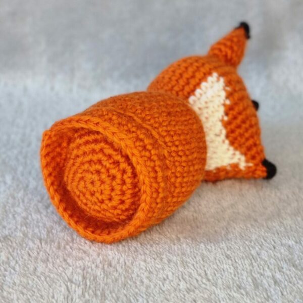 A crocheted orange fox soft toy lying on it's side. Close up of the round base of fox on a light grey background