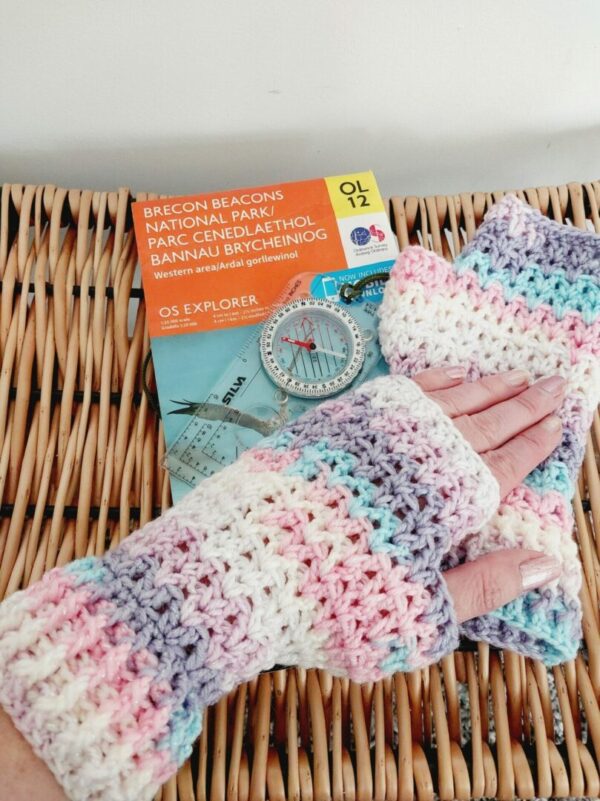 Crochet fingerless gloves, in an 'Unicorn' colourway of pastel pink, purple, blue and white. Shown on makers hand laying flat on top of a map and the 2nd mitt.