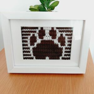 Crochet mosaic picture of a dog's paw in brown and white in a white frame.