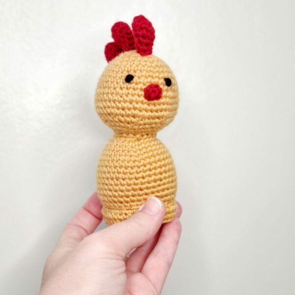 A crochet yellow and red chicken being held by a hand