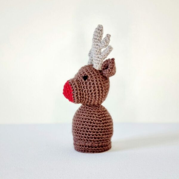 Side view of a crochet brown reindeer soft toy with a red nose on a white background