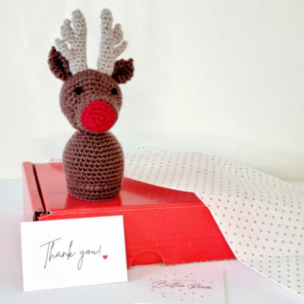 A brown reindeer soft toy with a red nose sat on top of red postage box with white packaging
