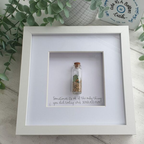 White box frame with small cork topped glass bottle containing sea treasures.