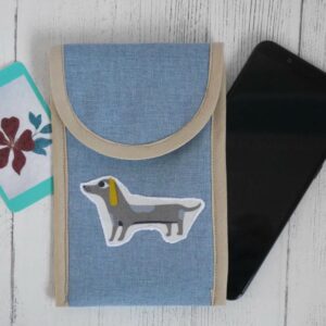 A pale blue-grey fabric phone case with a cartoon sausage dog appliqued to the front and trimmed in cream binding shown with a mobile phone.