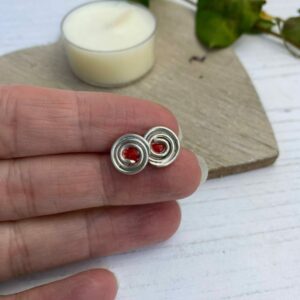 silver stud earrings with a swirl pattern and a red centre in fingers with wooden coaster and candle in the background