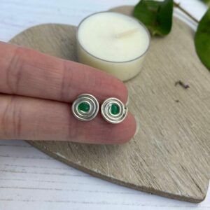 Silver swirl stud earrings with a green centre in between fingers with a wooden background and cream candle