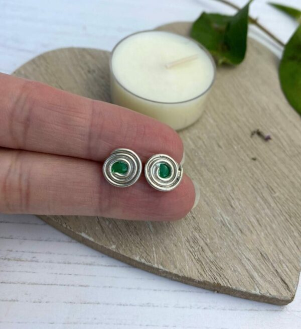 Silver swirl stud earrings with a green centre in between fingers with a wooden background and cream candle