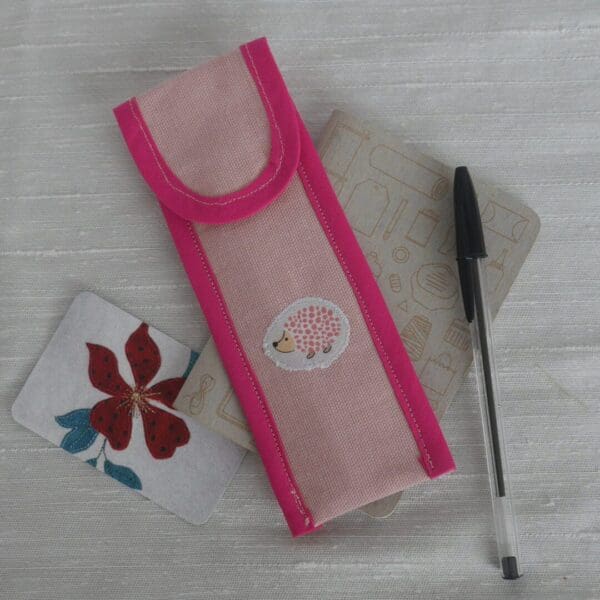 Pale pink rectangular pen case with bright pink trim and appliqued hedgehog on the front shown with pen and small notebook