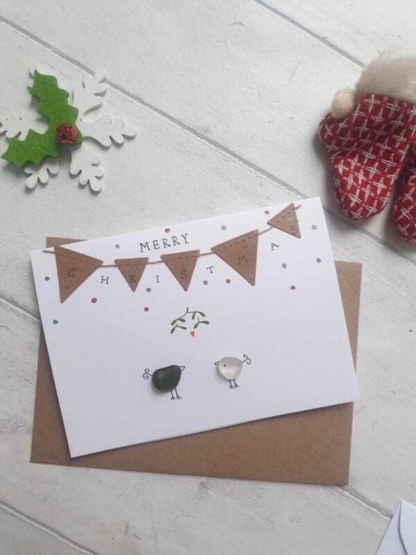 A6 greetings card, white. With Kraft bunting and Merry Christmas handwritten in black ink. Features 2 seaglass birds kissing underneath handdrawn mistletoe.