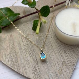 Gold necklace with swiss blue pear shaped faced topaz on wooden coaster with candle and leaves in the background