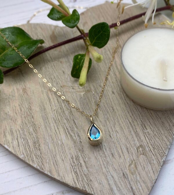 Gold necklace with swiss blue pear shaped faced topaz on wooden coaster with candle and leaves in the background