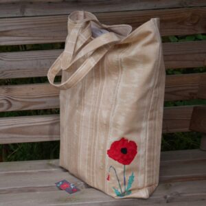 A beige tote bag with red poppy appliqued onto the lower right hand side of the front of the bag, shown full on wooden bench.