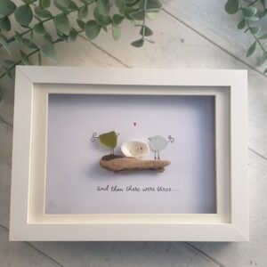 Two seaglass birds sitting on a piece of driftwood looking over a baby seaglass bird nestled in an open shell.