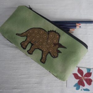 A green pencil case with an appliqued triceratops shown with pencils protruding from the zipped opening.