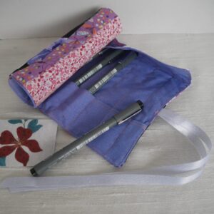 A patchwork roll shown partly closed with some pens in slots inside. The inner fabric is light purple the visible outer fabric is a pink and white print.