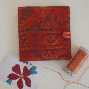 Closed needle case made from orange and purple damask fabric, shown with reel of thread and needle