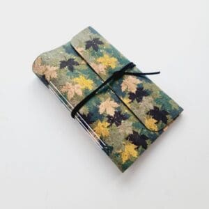 Cork fabric notebook journal with green and gold leaves pattern. Photographed standing again a green plant with silver pen in front.