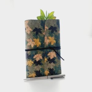 Cork fabric notebook journal with green and gold leaves pattern. Photographed standing again a green plant with silver pen in front.