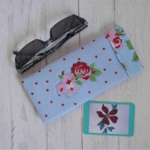 Light blue fabric glasses case with pink floral appliqued design shown with sunglasses.