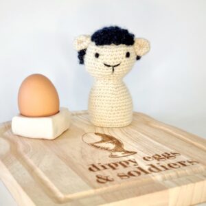 Black and white crochet seep sat on a wooden board with an egg cup and egg