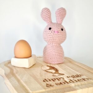 Pink crochet bunny rabbit egg cosy sat on a wooden board with a white egg cup and egg