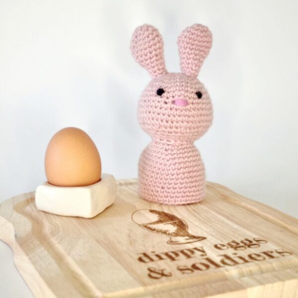 Pink crochet bunny rabbit egg cosy sat on a wooden board with a white egg cup and egg