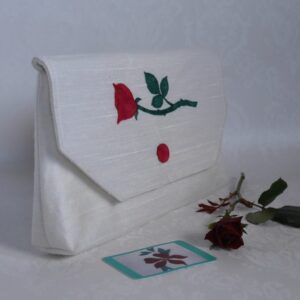 White textured fabric clutch bag with appliqued red rose on flap, shown with single red rose and business card.
