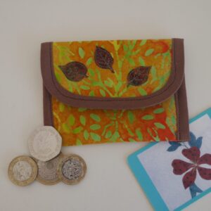 Coin purse made from batik printed fabric in shades of yellow, orange and green, trimmed in brown and with three appliqued brown leaves on the flap. Shown with a selection of coins and a business card.