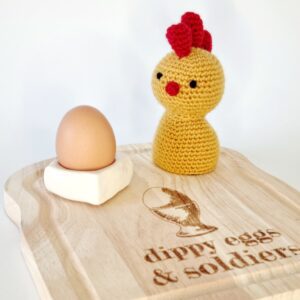 Yellow crochet chicken egg cosy say on a board with a egg cup