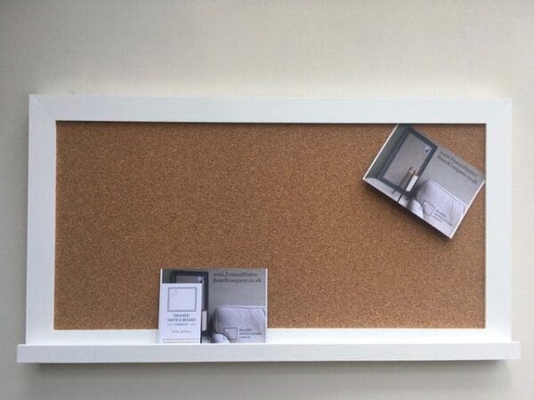 A large cork pin board with modern white frame and shelf hangs on a neutral wall.