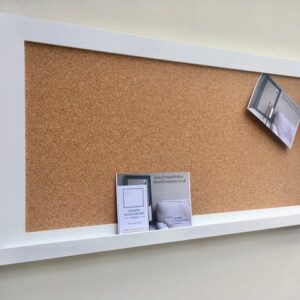A rectangular cork pin with modern white frame and white shelf hangs on a neutral wall.