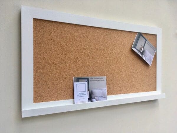 A rectangular cork pin with modern white frame and white shelf hangs on a neutral wall.