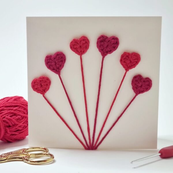 Square cream card with hearts on surrounded by a ball of yarn, scissors and crochet hook