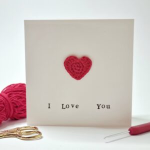 A square cream greetings card with the words 'I love you' written on the front and a red heart. Surrounding the card is a ball of red yarn, gold scissors and a crochet hook. All on a white background