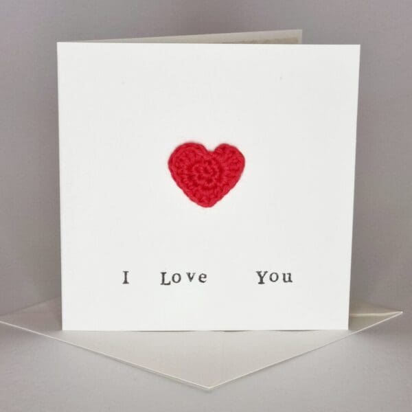 A simple cream card with the words 'I love you' written on the front and a red love heart. It's sat on top of a cream envelope. All on a white grey background