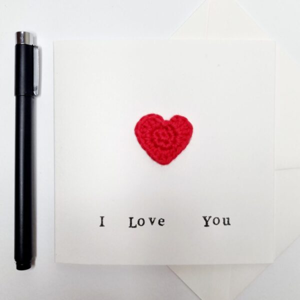 A white card with a red crochet heart on the front and the words 'I love you' written on the front in black ink. Lying next to a black pen