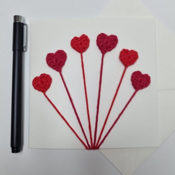 A square cream greetings card with 6 red hearts on the front next to a black pen