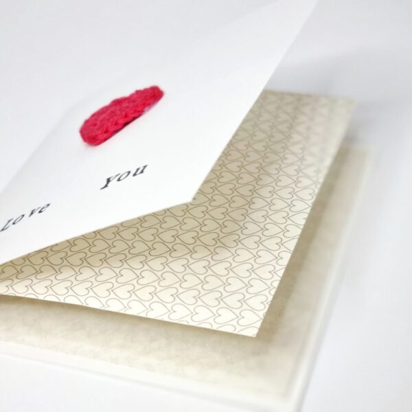 A greetings card lying flat on a white surface with a red heart on the front and a paper insert with a gold heart pattern on.