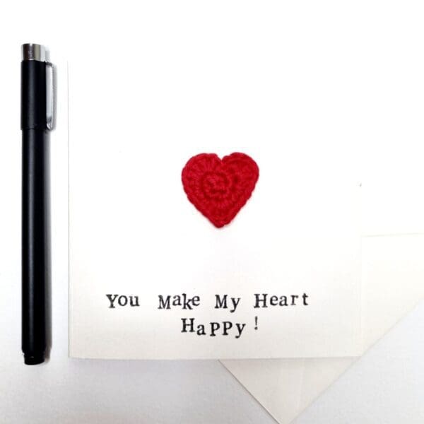 A white greetings card with the words 'you make my heart happy' written on the front with a red crochet heart. A black pen lined up beside it