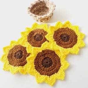 Four yellow and brown crochet sunflower coasters made by Sarah Lou Crafts shown laying together on a white table with brown crochet storage pot above.