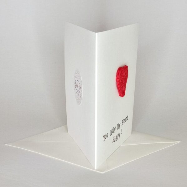 A side view of a white greetings card on a white background with a red crochet heart on the front.