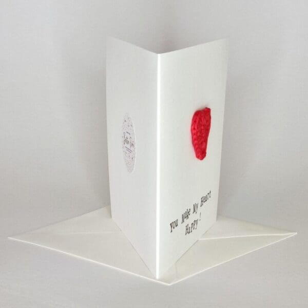 A side view of a white greetings card on a white background with a red crochet heart on the front.